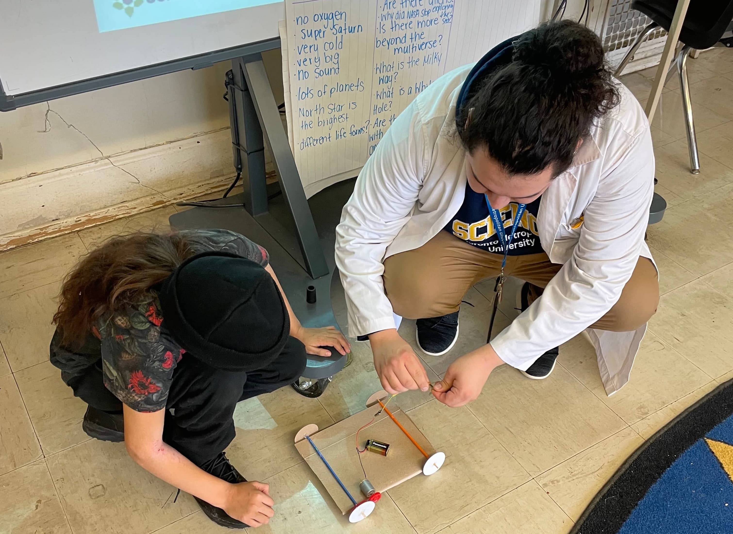 Caleb Wesley kneeling down working with a student on fixing their DC motor car during an in class workshop.